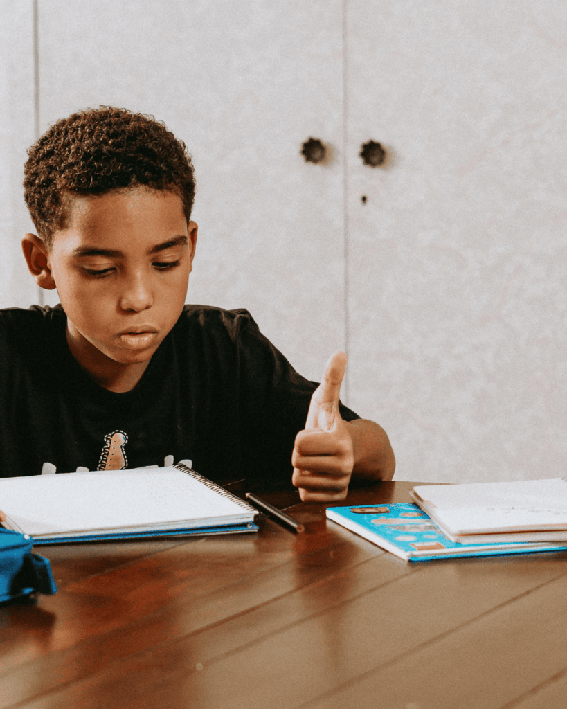 Imagem de um menino negro sentado que está estudando com um lápis na mão e caderno na mesa.